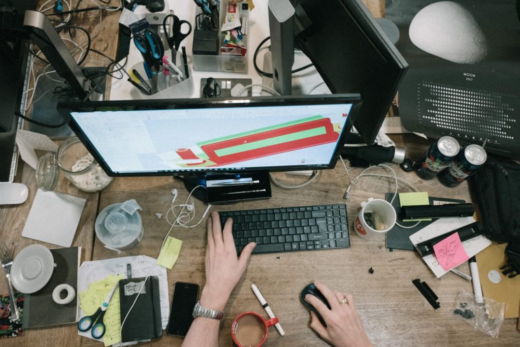 Top view of a cluttered workspace with a computer monitor, keyboard, notes, coffee cups
